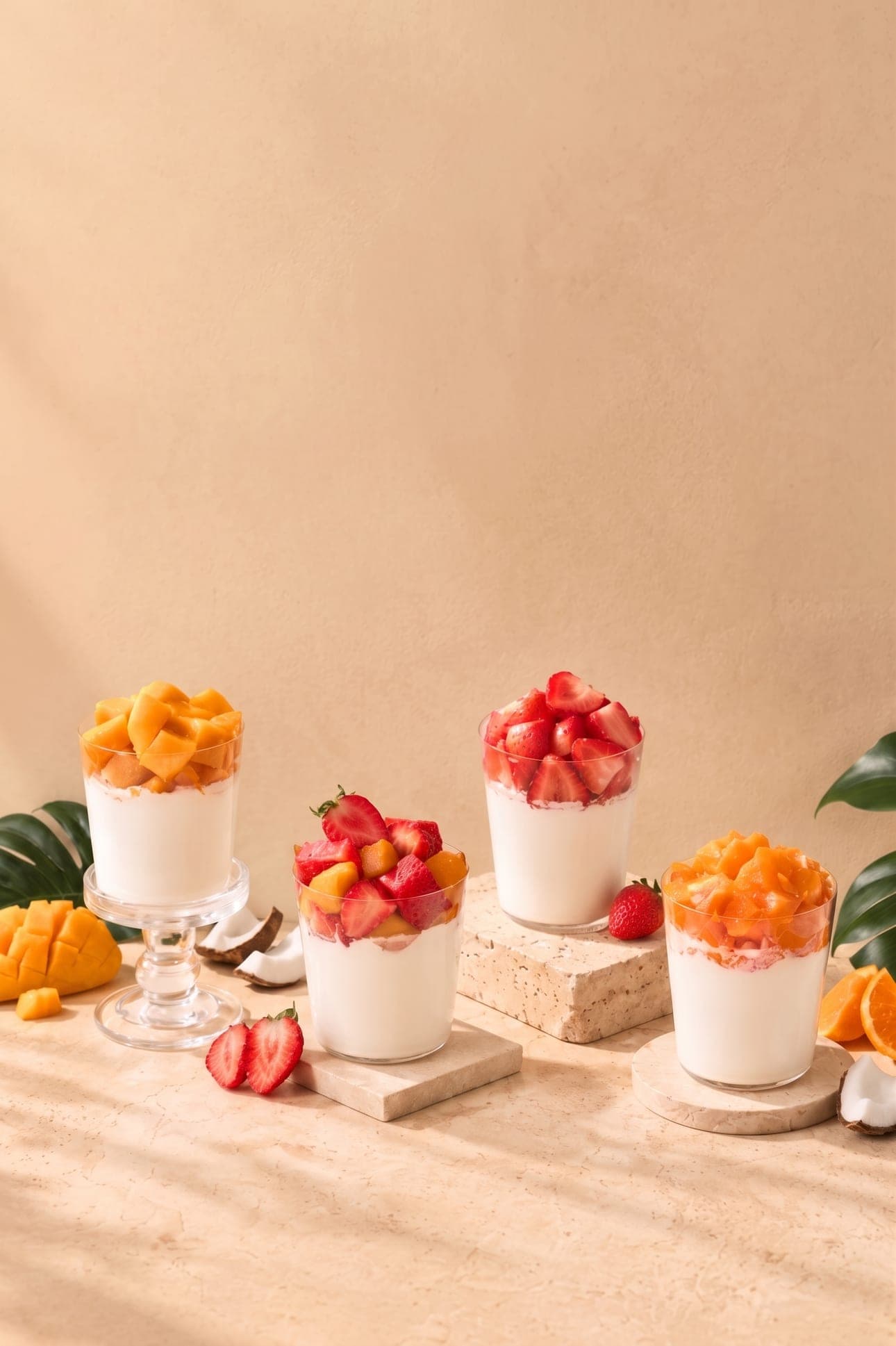Four coconut pudding glasses topped with mango, strawberry and orange on travertine risers, against a warm beige backdrop.