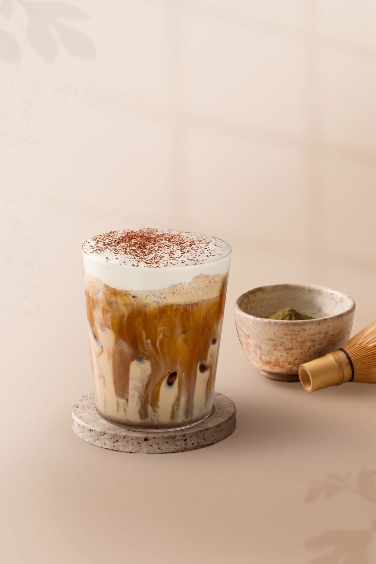 An iced creamy hojicha latte dusted with cocoa on a stone coaster, beside a bowl of hojicha powder and a bamboo whisk.