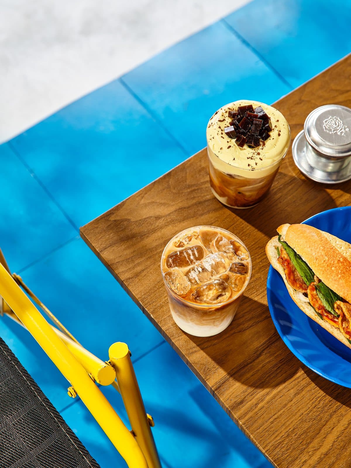 A bánh mì and two iced Trạm coffees on a wooden board, on a blue table